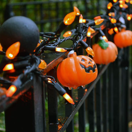 Festive Halloween decorations featuring a string of orange lights and small plastic pumpkin lanterns with jack-o'-lantern faces, wrapped around a black metal fence, creating a spooky and bright atmosphere.