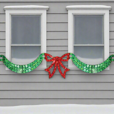 This is a lifestyle image showing the 6 foot long lighted Christmas decoration designed to look like green ribbon with a red bow in the center. In this image, it's across two windows at the front of a snowy house.