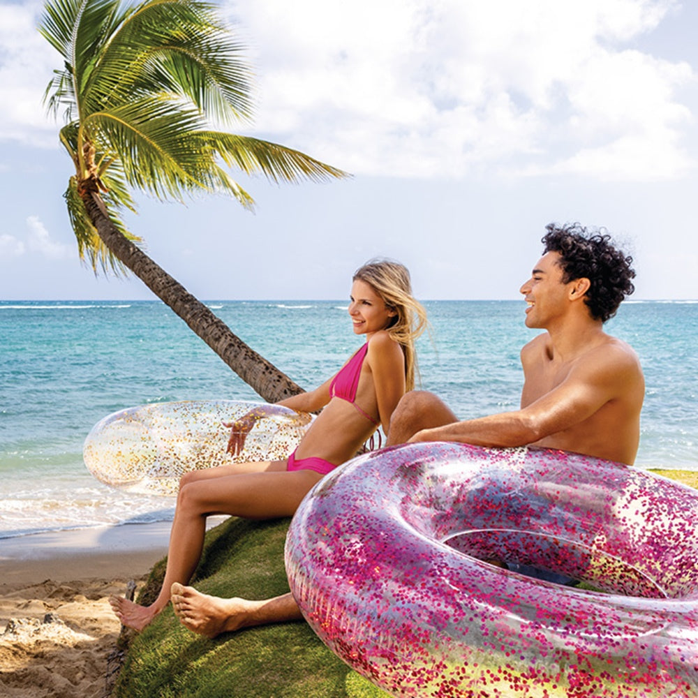 This is a lifestyle image showing a woman and a man sitting on a beach holding two of these glitter inner tubes.