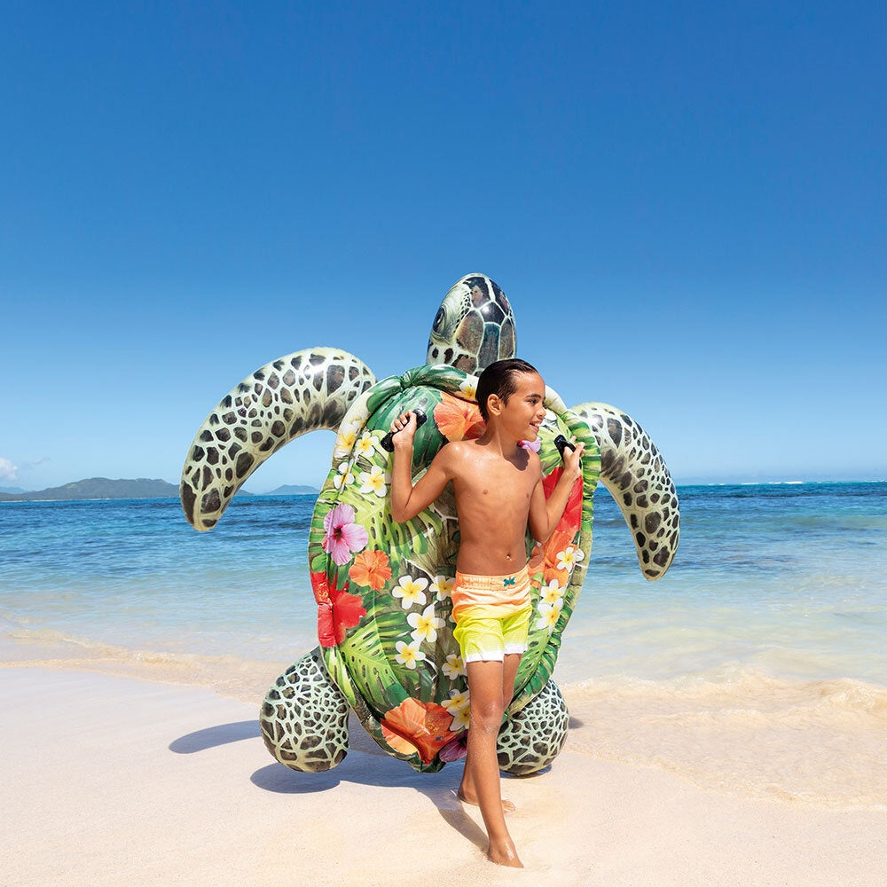 This is a lifestyle image showing a young boy standing on the beach, holding the turtle inflatable.