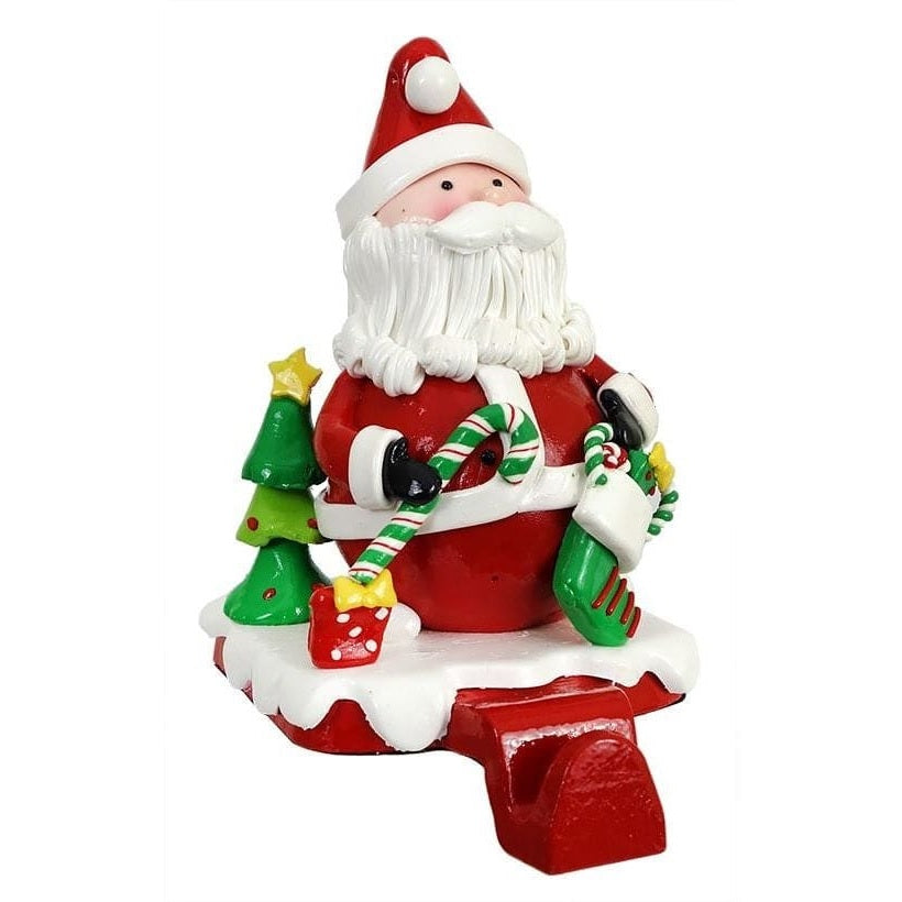 Stocking holder with Santa on it. He's holding a candy cane and a stocking. He's standing next to a small decorated Christmas tree and a present.