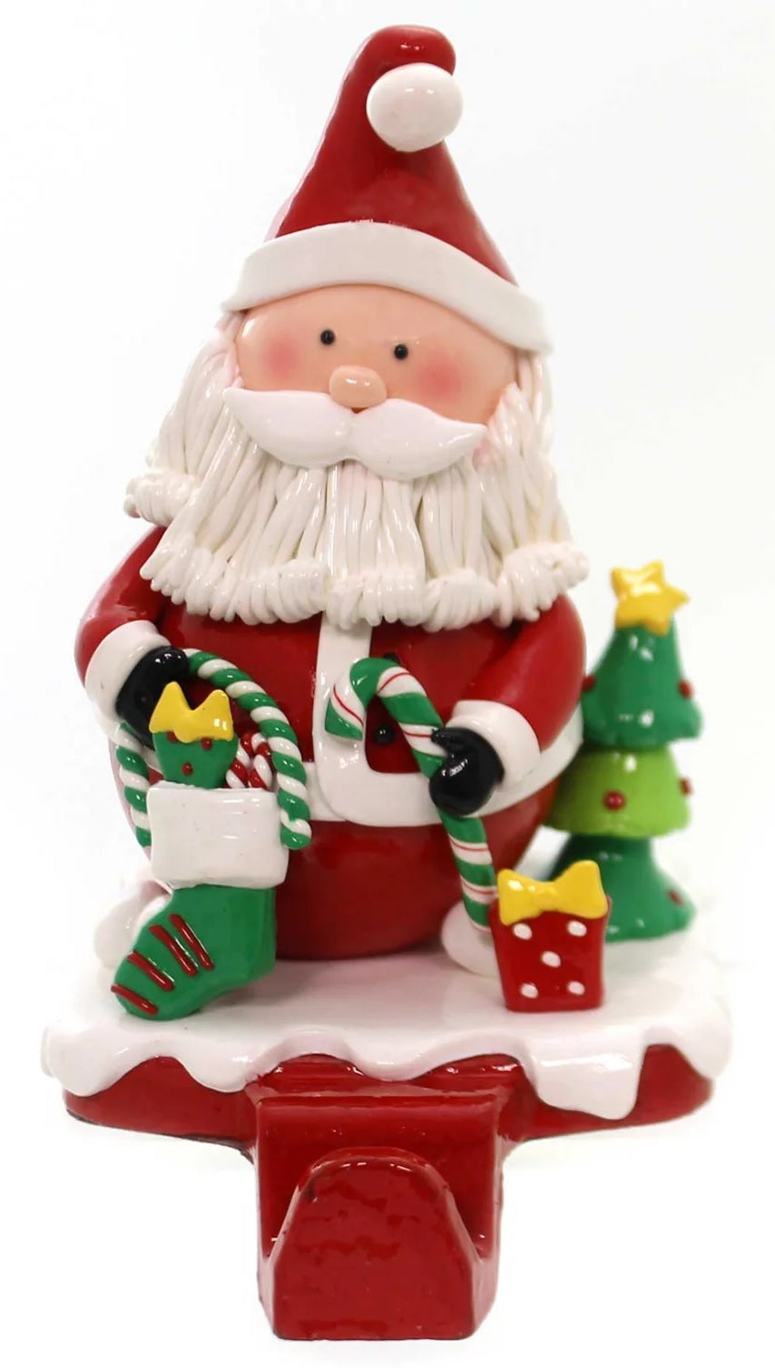 Stocking holder with Santa on it. He's holding a candy cane and a stocking. He's standing next to a small decorated Christmas tree and a present.