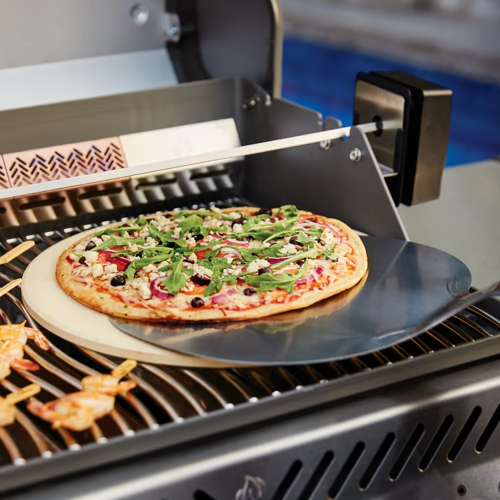 This is a lifestyle image showing a pizza stone on a grill. There's a pizza on the circular spatula and it's being slid onto the pizza stone.