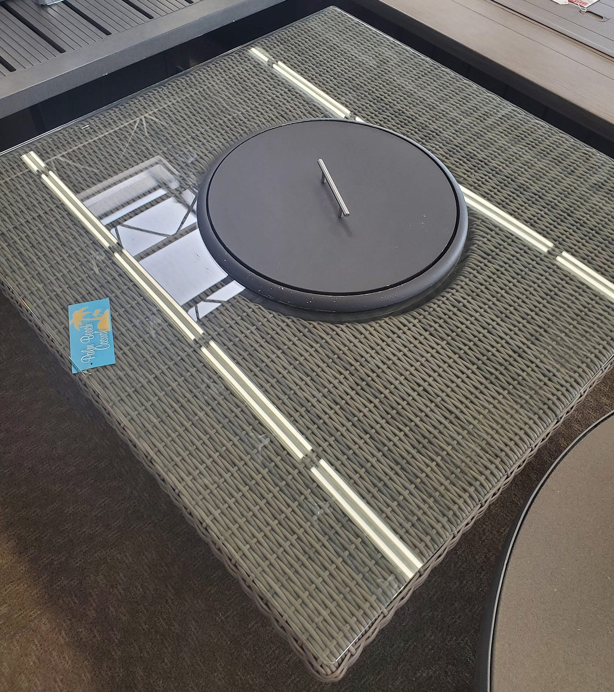 Square fire pit table. Made of woven materials. Features a lid. Underneath the lid is fire glass.