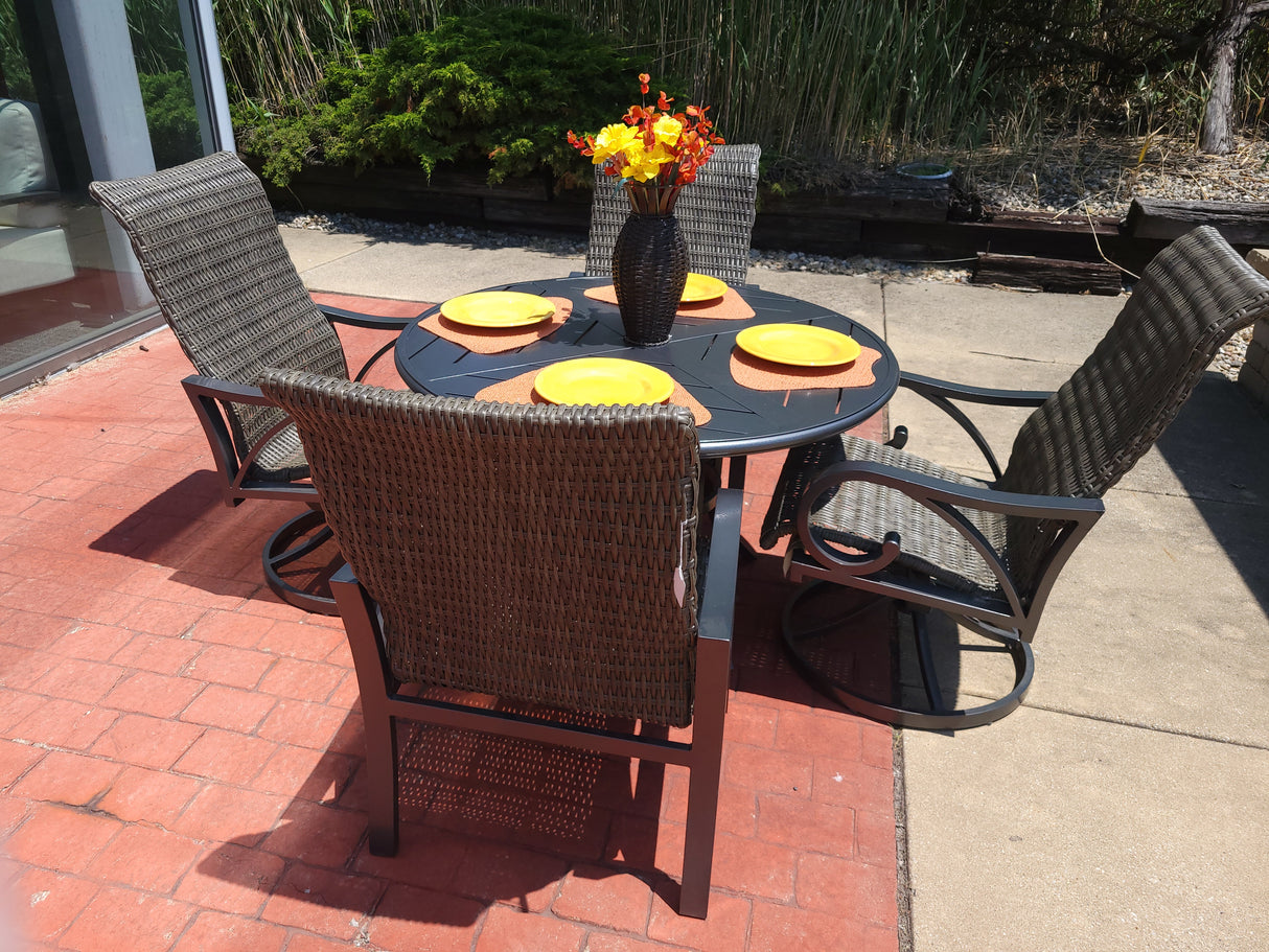 This image shows a brown wicker patio dining set with a round brown table in the center. There are four chairs. On the table, there are four yellow plates on four orange mats. In the center of the plates, there's a vase with yellow and red flowers inside.