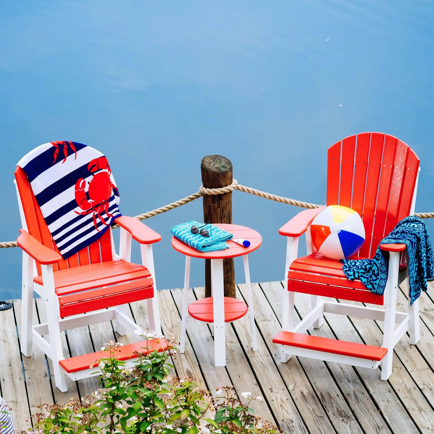 This is a lifestyle image showing two red and white colored Adirondack chairs and a matching end table between them. They're all set up on a pier next to a body of water. The chair on the left has a blue and white striped towel draped over it. On the towel is a red crab design. On top of the end table, there's a folded blue shawl, some sunglasses and a tube of suntan lotion. On the chair on the right, there's an inflatable beach ball and a blue bathing suit cover up.