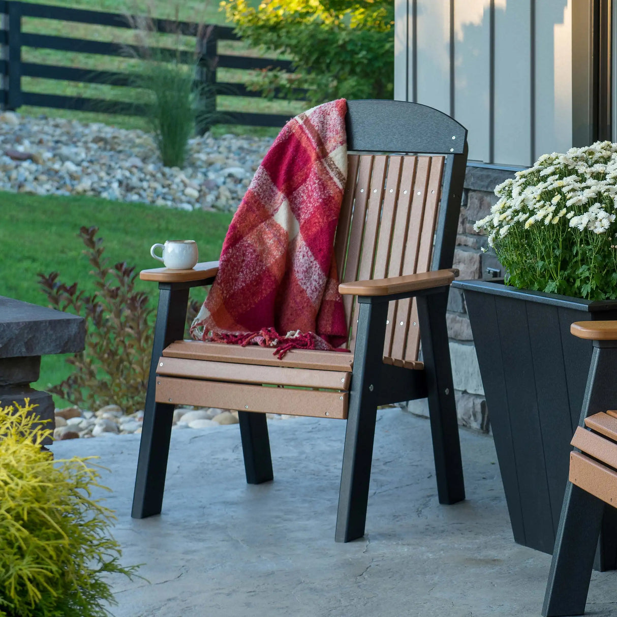 This is a lifestyle image showing the Cedar and Black colored bench on a stone patio with a red blanket draped over the side and a mug on the armrest.