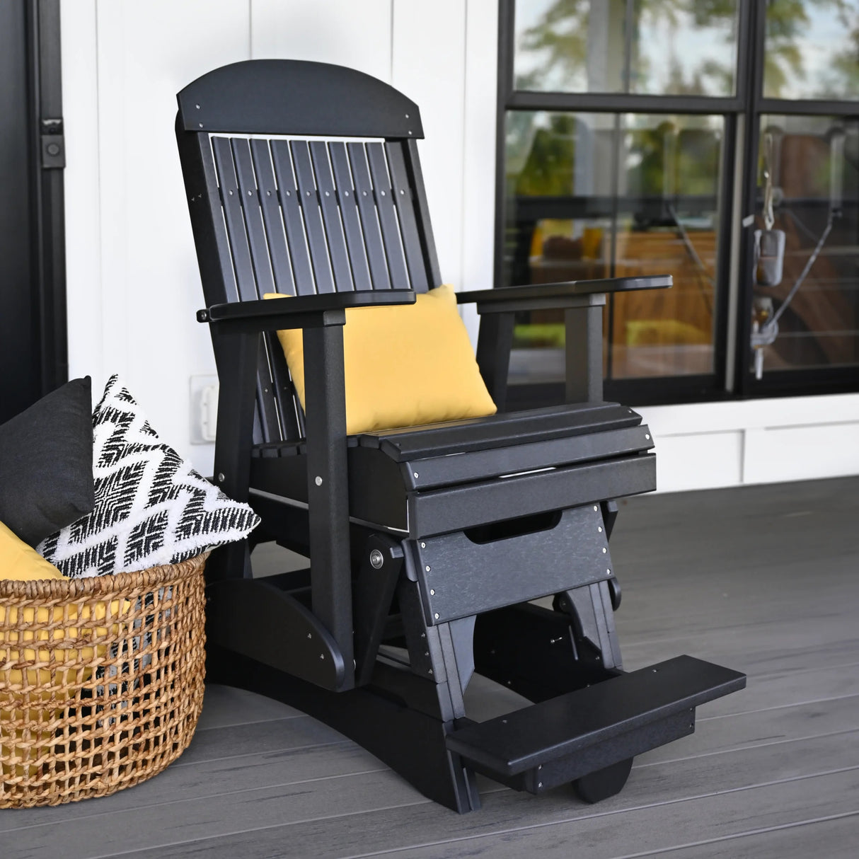 This is the lifestyle image of the Black colored balcony glider chair on a patio in front of a house. There's a yellow throw pillow on the seat.