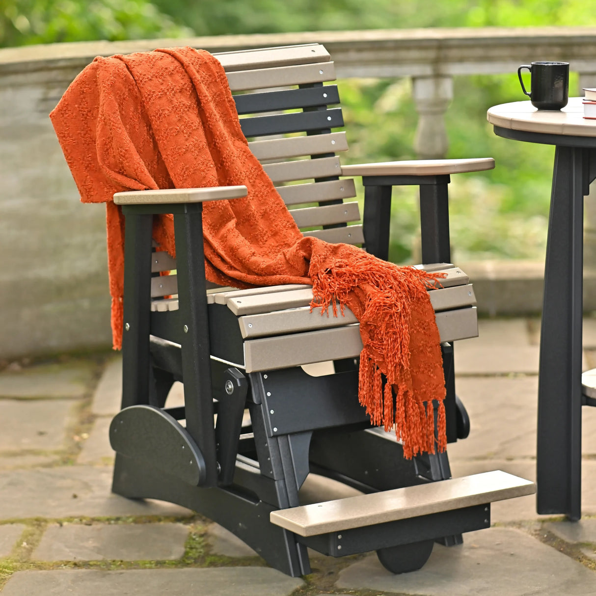 This is a lifestyle image showing the Weatherwood and Black colored glider chair with an orange blanket draped over the side.