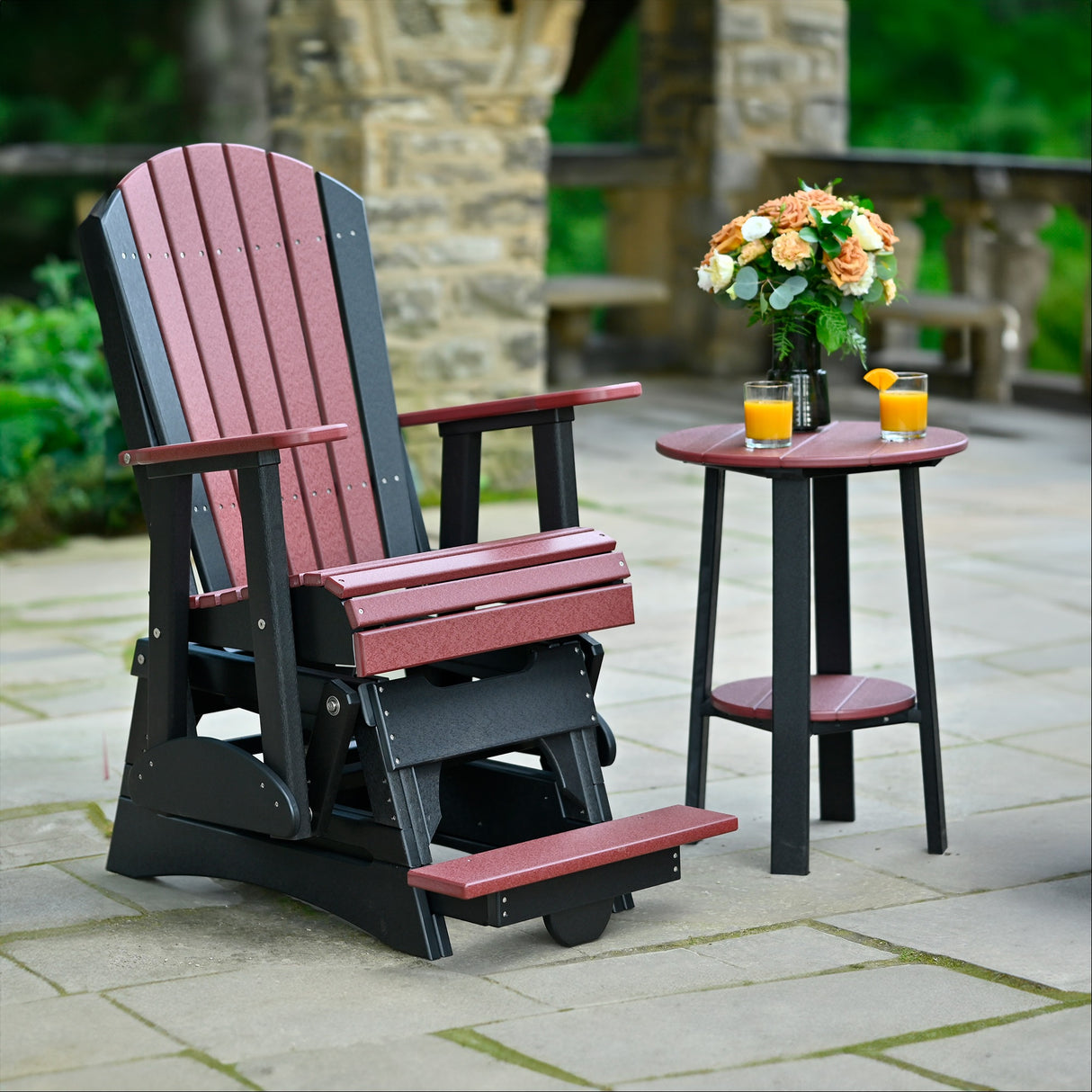 This is a lifestyle image showing the Cherrywood and Black colored Adirondack glider chair on a stone patio.