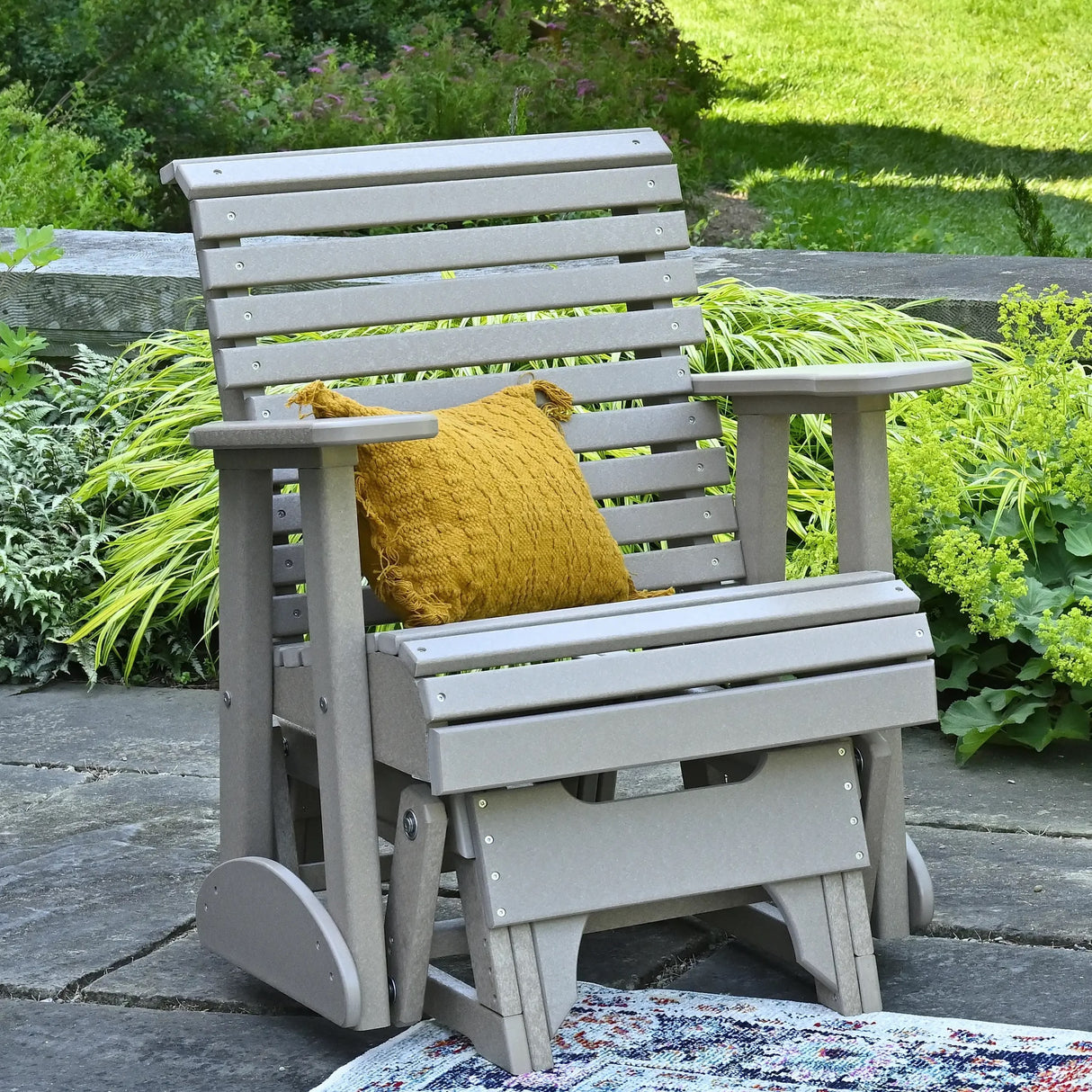 This is a lifestyle image showing off the Weatherwood glider chair on a stone patio. There's a yellow throw pillow on the seat.
