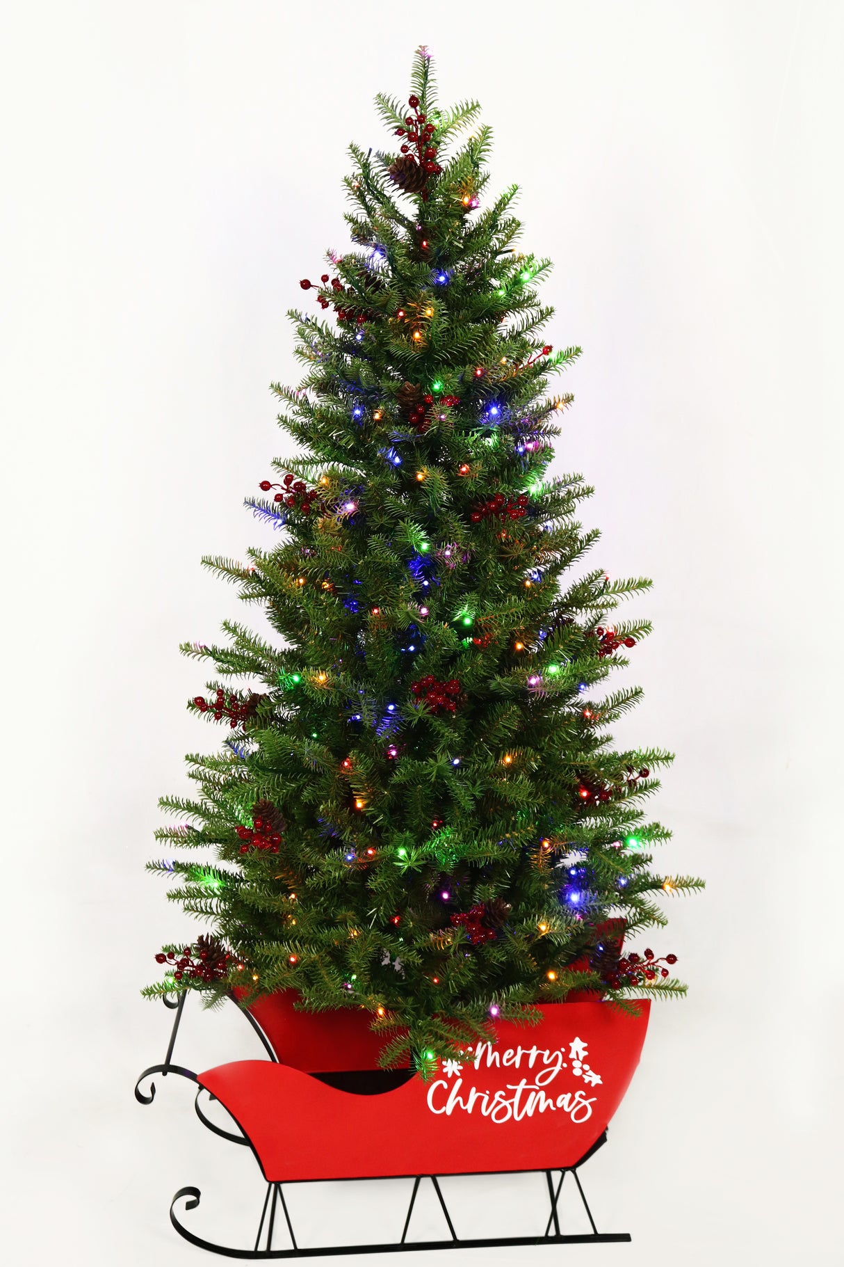 Pre-lit artificial tree in a red sleigh. The branches feature red berries and the red sleigh reads "Merry Christmas" in white, cursive font.