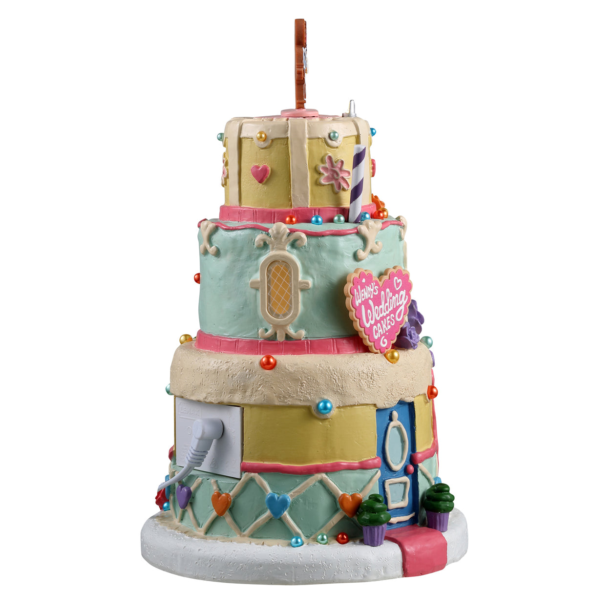 Resin lighted building for a Lemax Village display. Designed to look like a three tiered wedding cake. There's a gingerbread cookie couple on top and a cookie-shaped sign that reads "Wendy's Wedding Cakes." The top tier is yellow, the middle tier is a light teal color and the bottom tier is both yellow and light teal. There are sprinkles and frosting accents all over.