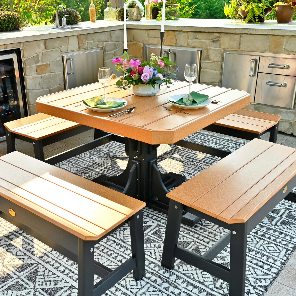 This is a lifestyle image showing the Cedar and Black colored square dining table surrounded by matching benches. The table is surrounded by a stone outdoor kitchen island.