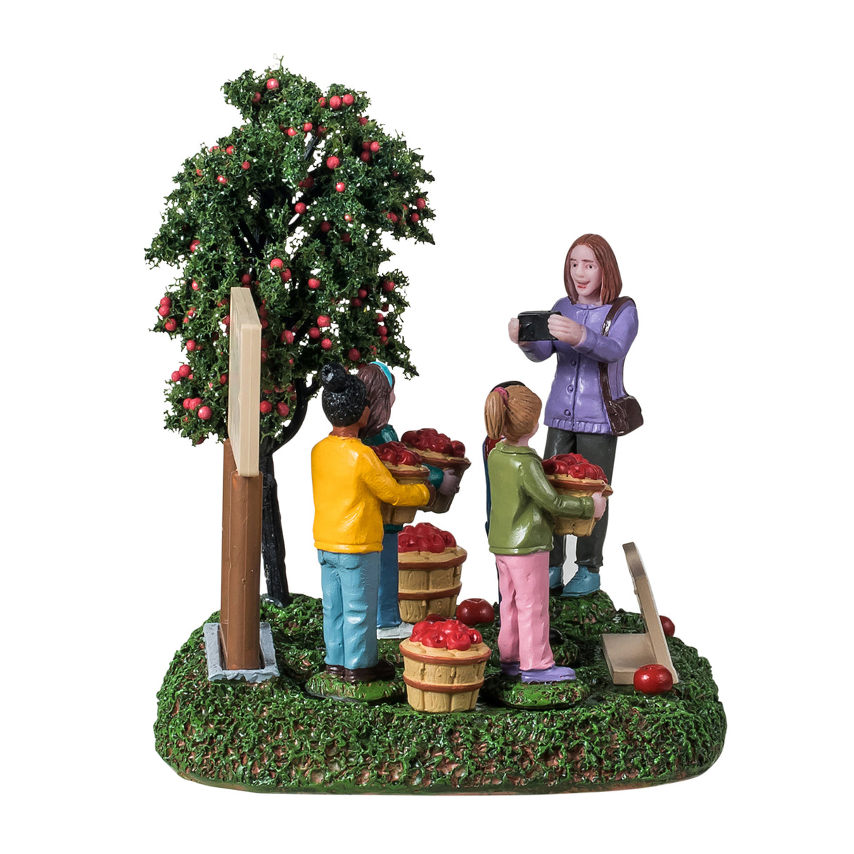 Resin table accent for a Lemax Village tabletop display. Designed to look like four students posing for a picture. They're standing next to an apple tree and a sign that reads "Annie's Apple Orchard. Pick your own farm fresh apples." The children are all smiling and holding a basket of apples. There are apples on the ground and a sign that reads "Park Hill Elementary - Mrs. Johnson's class." Mrs. Johnson is the one taking a picture of the students. The ground is covered in grass.