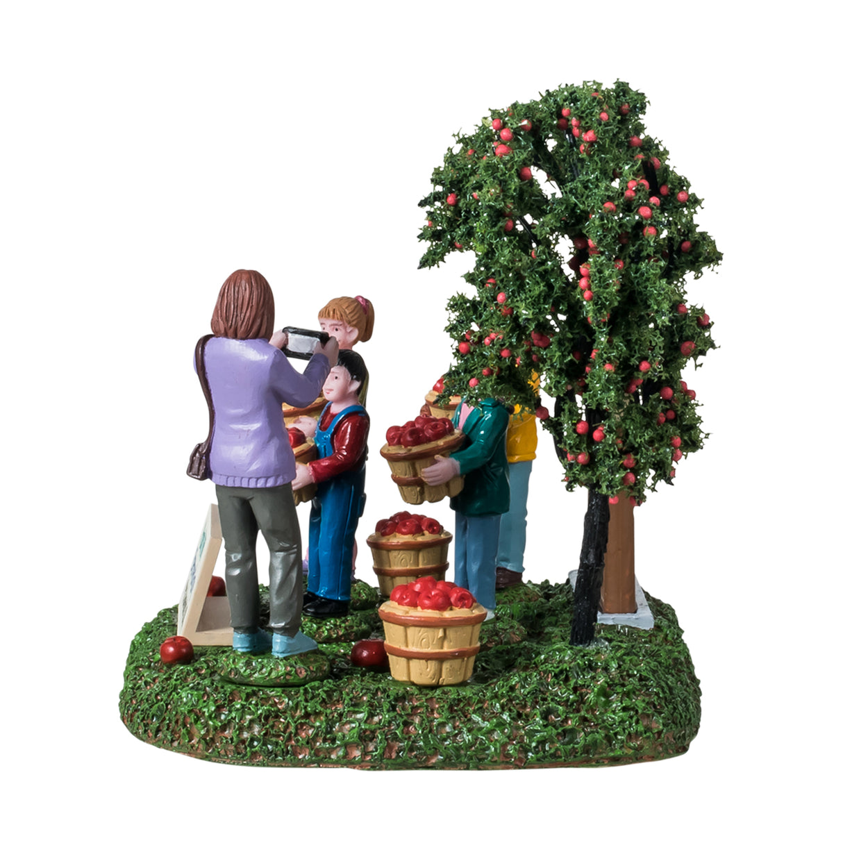 Resin table accent for a Lemax Village tabletop display. Designed to look like four students posing for a picture. They're standing next to an apple tree and a sign that reads "Annie's Apple Orchard. Pick your own farm fresh apples." The children are all smiling and holding a basket of apples. There are apples on the ground and a sign that reads "Park Hill Elementary - Mrs. Johnson's class." Mrs. Johnson is the one taking a picture of the students. The ground is covered in grass.