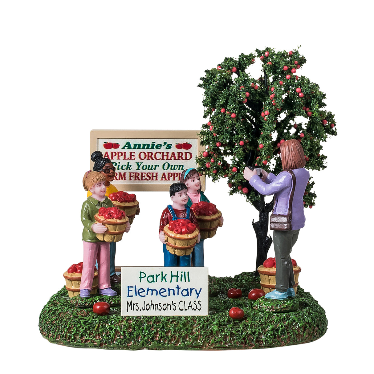 Resin table accent for a Lemax Village tabletop display. Designed to look like four students posing for a picture. They're standing next to an apple tree and a sign that reads "Annie's Apple Orchard. Pick your own farm fresh apples." The children are all smiling and holding a basket of apples. There are apples on the ground and a sign that reads "Park Hill Elementary - Mrs. Johnson's class." Mrs. Johnson is the one taking a picture of the students. The ground is covered in grass.