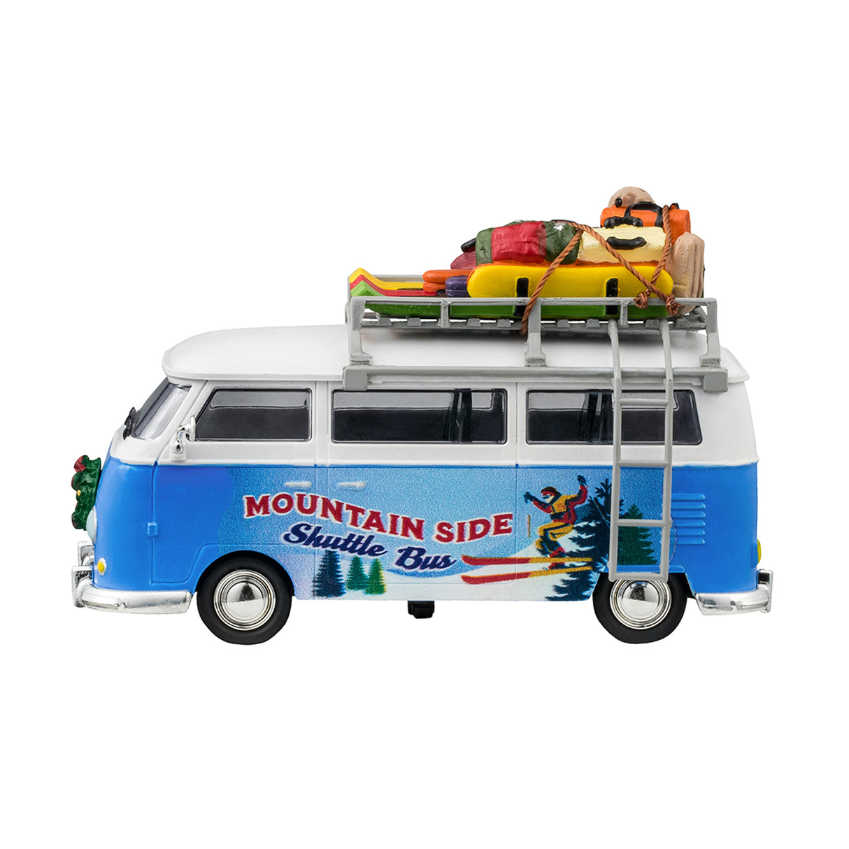 Plastic table accent for a Lemax display. Designed to look like a blue and white Volkswagen shuttle bus. The sides of the bus read "Mountain Side Shuttle Bus" and features a mural of someone skiing down a snowy hill surrounded by trees. There's a wreath on the front bumper. There's a ladder on the side leading up to the roof o the bus that's holding a bunch of ski equipment and luggage.