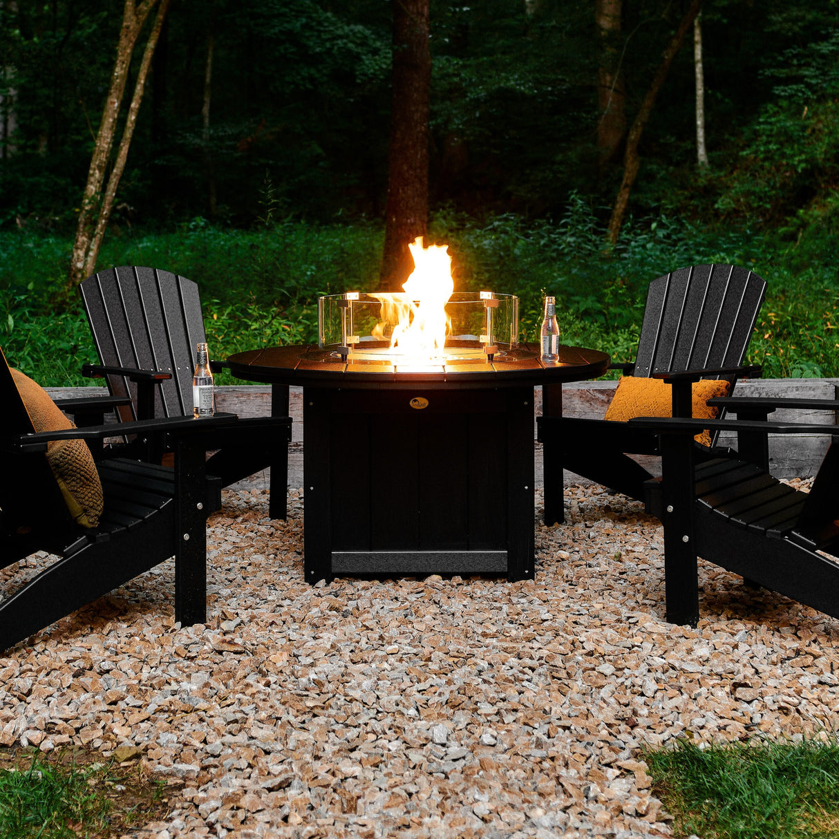 This is a lifestyle image showing the Black colored fire pit table surrounded by Black colored Adirondack chairs. There's a fire inside the table.