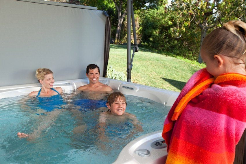 This image shows a mom, a dad and a young boy inside a hot tub smiling at a young girl who is using a towel to dry off outside of the hot tub. They used the lifter system to move the hot tub cover into an upright position to use the cover as a privacy barrier.