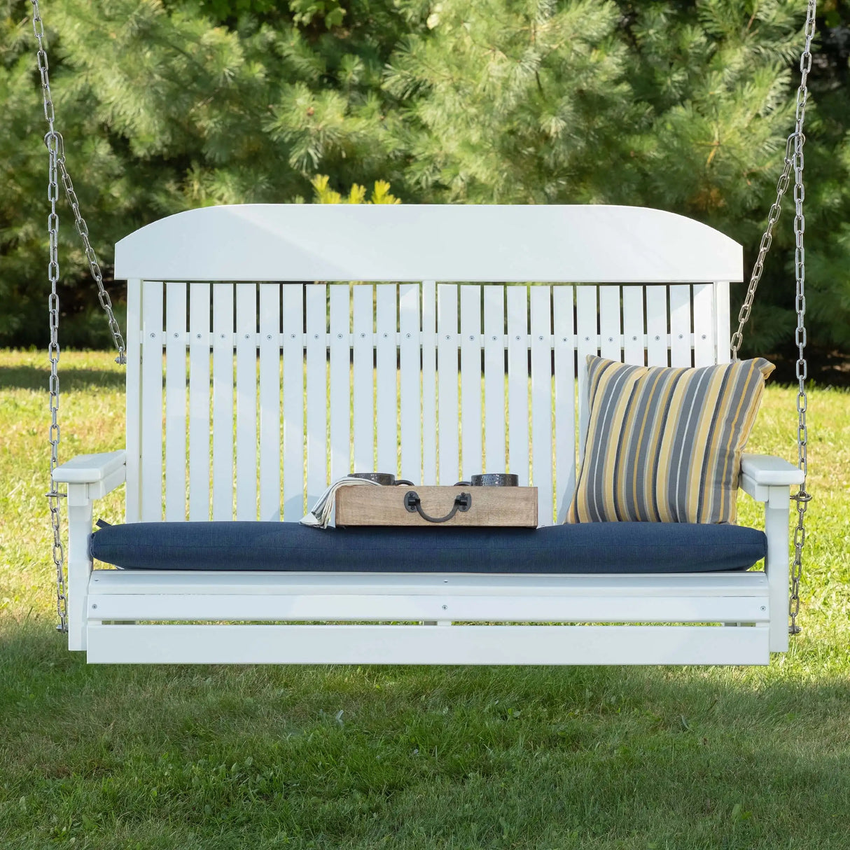 This is a lifestyle image showing the White swing in a grassy backyard. There's a blue seat cushion and a yellow and blue striped throw pillow.