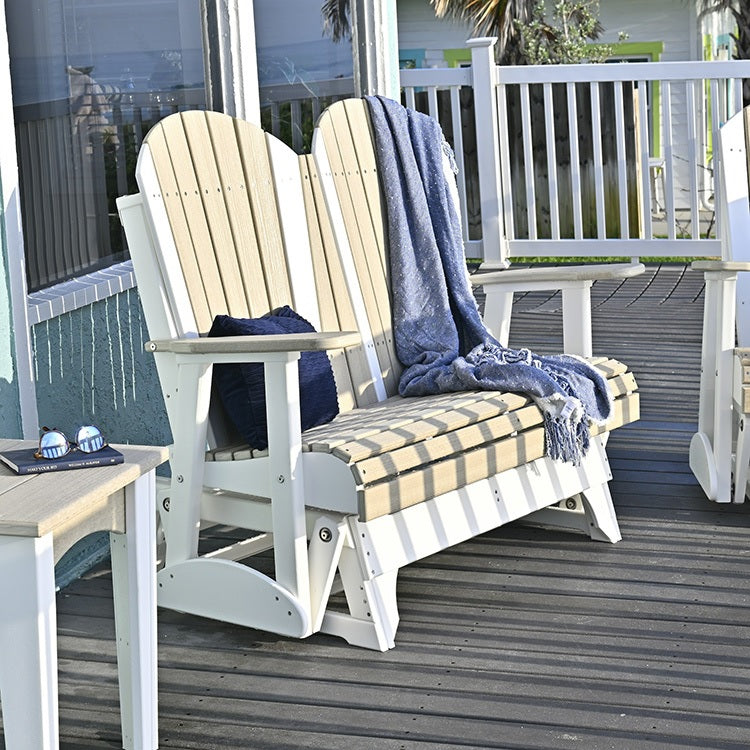 This is a lifestyle image showing the Birch and White Adirondack glider bench on a wooden porch. There's a light blue blanket draped over the side and a dark blue throw pillow on the left side.