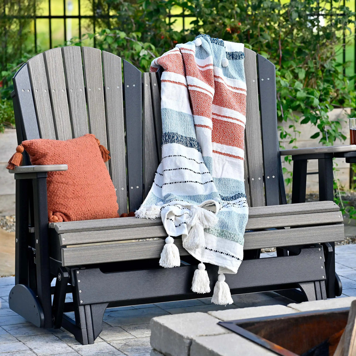 This is a lifestyle image showing the Weatherwood and Black colored Adirondack glider bench. There's an orange throw pillow on the side and a white, orange and blue colored blanket draped over the side.