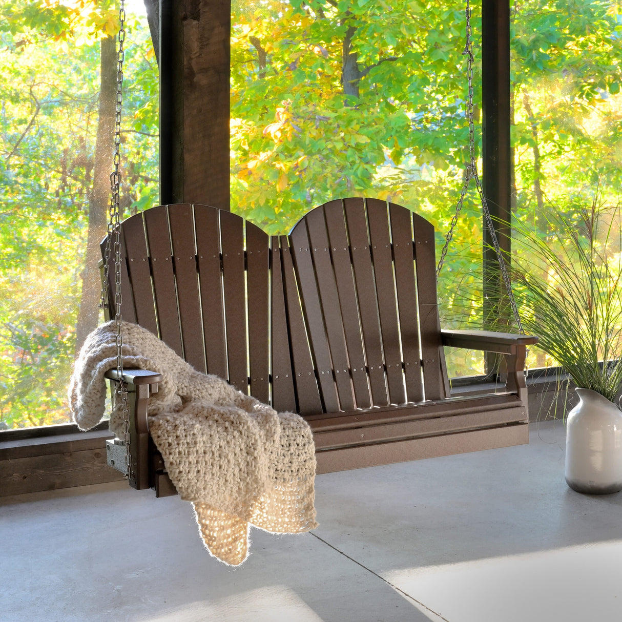 This is a lifestyle image showing off the Chestnut Brown colored Adirondack porch swing. It's inside a sun room and you can see a bunch of trees through the glass in the background.