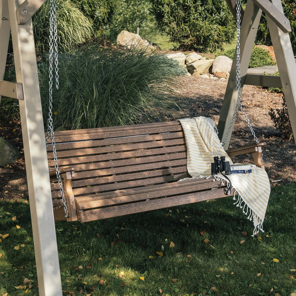 This is a lifestyle image showing the Antique Mahogany porch swing in a grassy backyard. There's a yellow and white striped blanket draped over the side and a pair of binoculars resting on the seat.