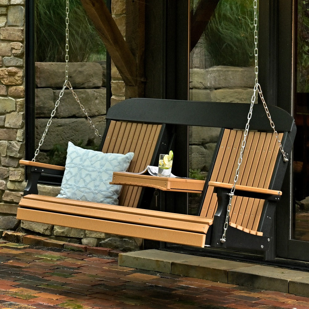 This is a lifestyle image showing the Cedar and Black porch swing in front of a home. There's a blue throw pillow on one side and a drink inside one of the cupholders.