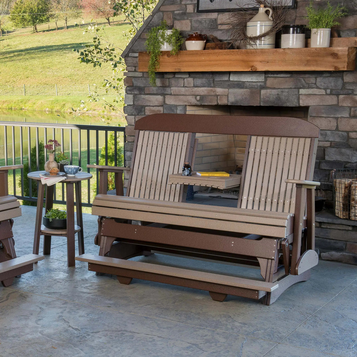 This is a lifestyle image showing the Weatherwood and Chestnut Brown colored balcony glider bench on a patio in front of an outdoor fireplace.