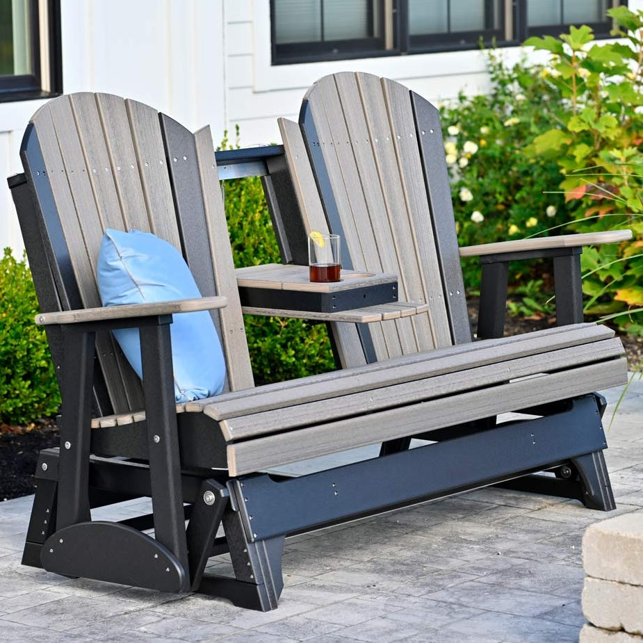 This is a lifestyle image showing the Weatherwood and Black colored Adirondack glider bench on a stone patio. There's a blue throw pillow on the side and the center console is down. There's a glass of juice in one of the cupholders.