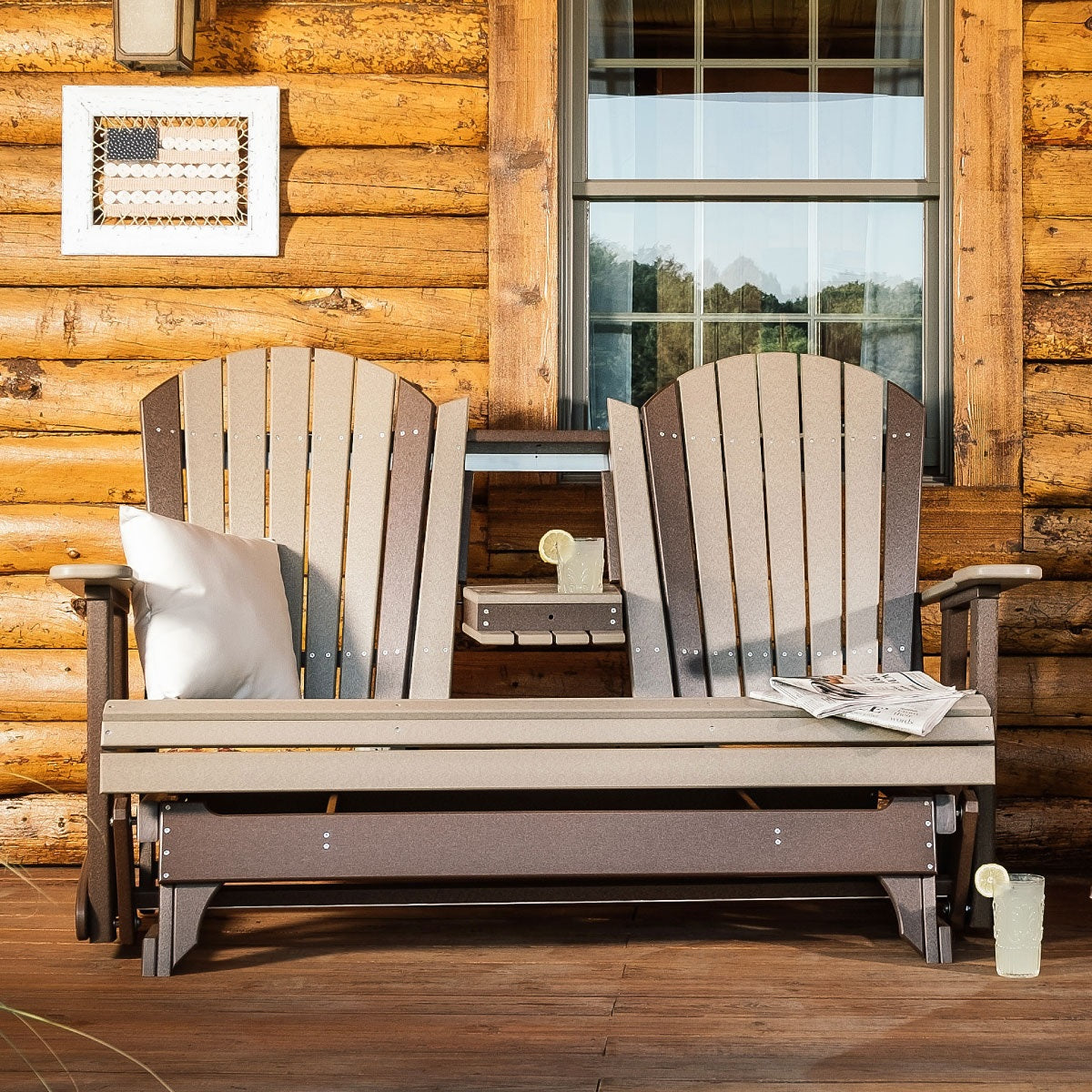This is a lifestyle image of the Weatherwood and Chestnut Brown colored Adirondack glider bench in front of a cabin.