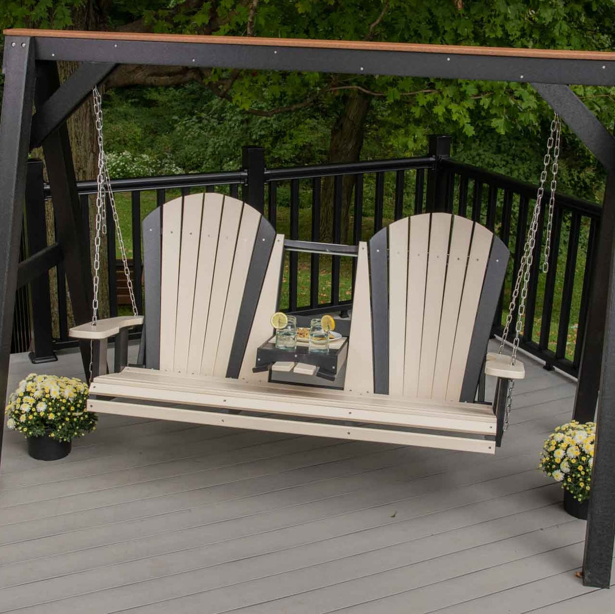This is a lifestyle image showing the Weatherwood and Black Adirondack swing on a wooden porch. The center console is down and there are two drinks in the cupholders.