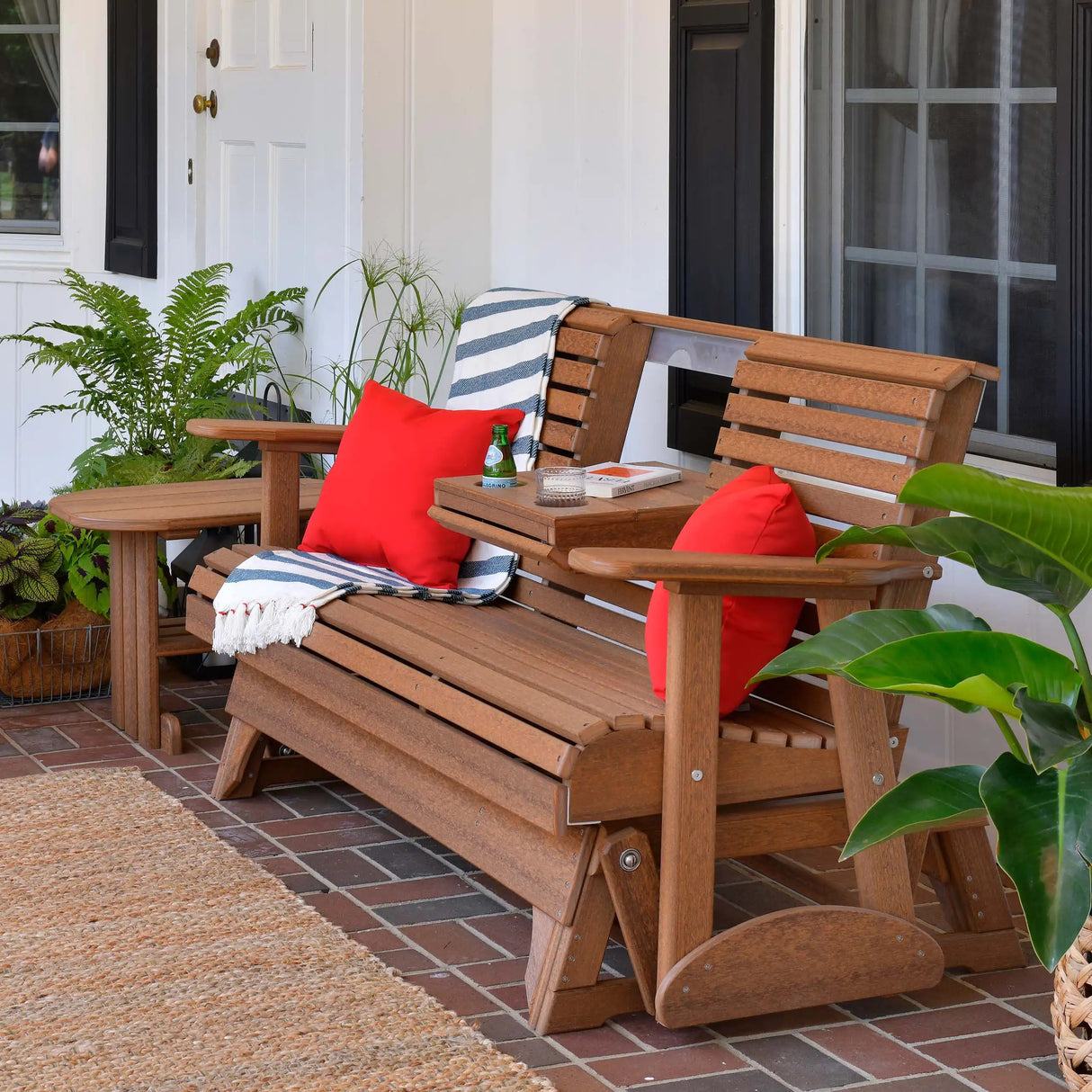 This is the Antique Mahogany colored glider bench on a brick patio. There are two red throw pillows on the sides and the center console is down. There's a bottle of sparkling water in one of the cupholders.