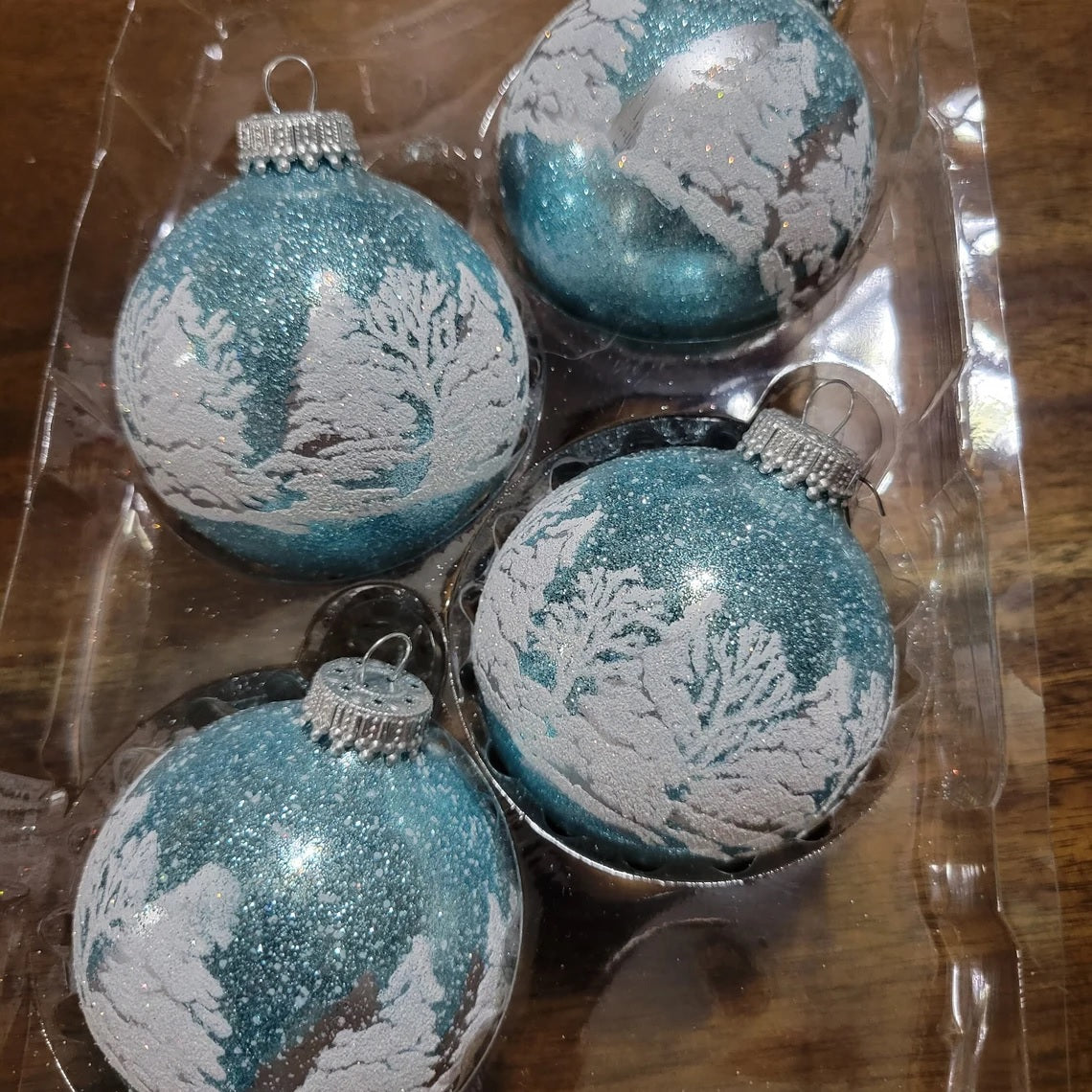 Set of ornaments decorated with light blue and white glitter. The ornaments have woodland scenes involving trees and a deer.