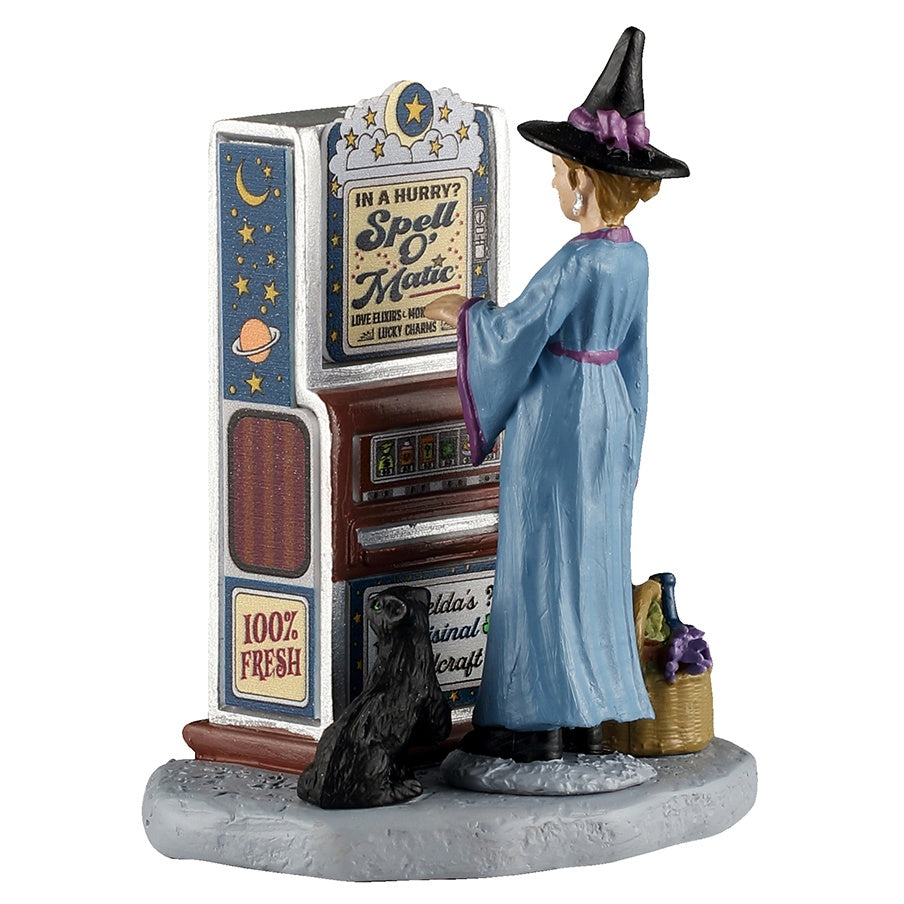 This image shows what this Lemax Spooky Town figurine looks like on a white background. It's designed to look like a witch wearing a light blue robe and a black, pointy hat standing in front of a vending machine. It reads "In a hurry? Spell O' Matic. Love elixirs and lucky charms. 100% fresh." Next to the witch, there's a small black cat with green eyes. On the other side of the witch, there's a basket of ingredients.