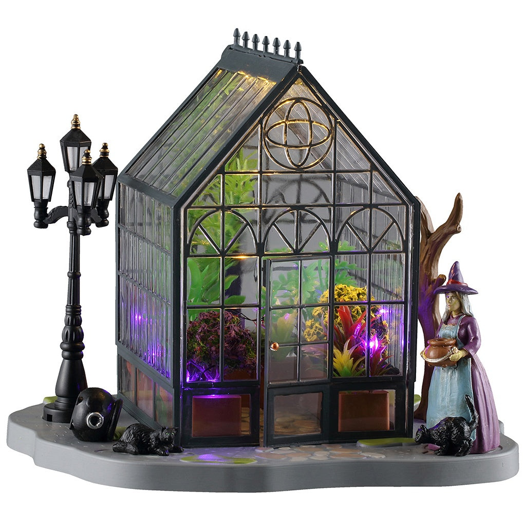 This image shows what this Lemax Spooky Town table accent looks like on a white background. It was designed to look like a greenhouse with plants inside and a witch standing outside.