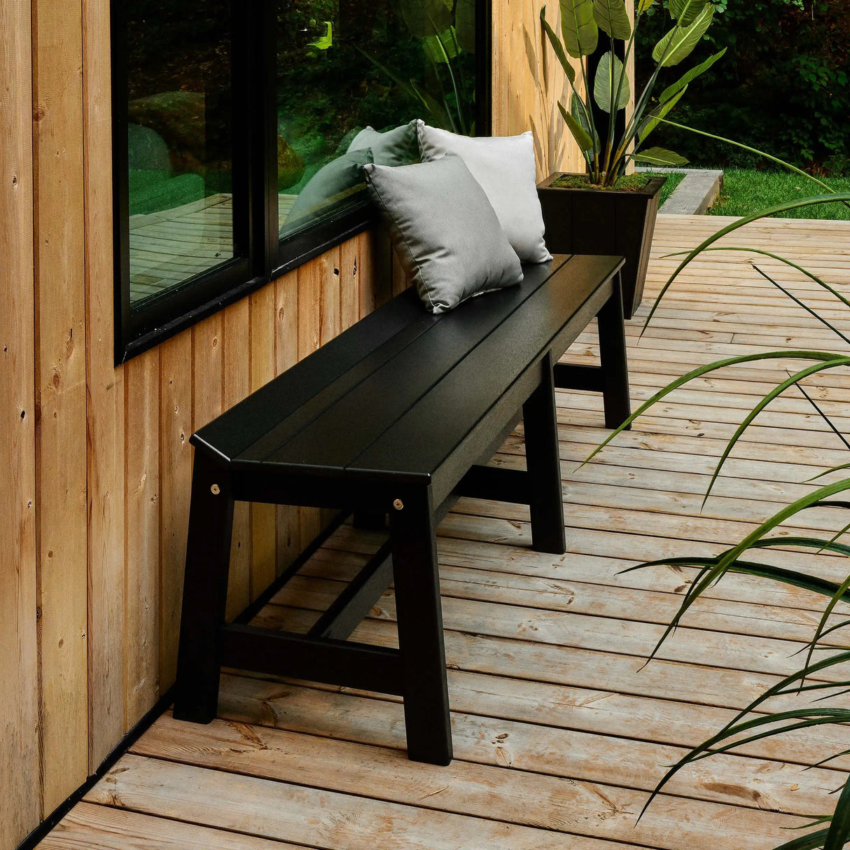 This is a lifestyle image showing the Black dining bench in front of a wooden house with a black window. There are two throw pillows placed on the bench: one is dark gray and the other is light gray.