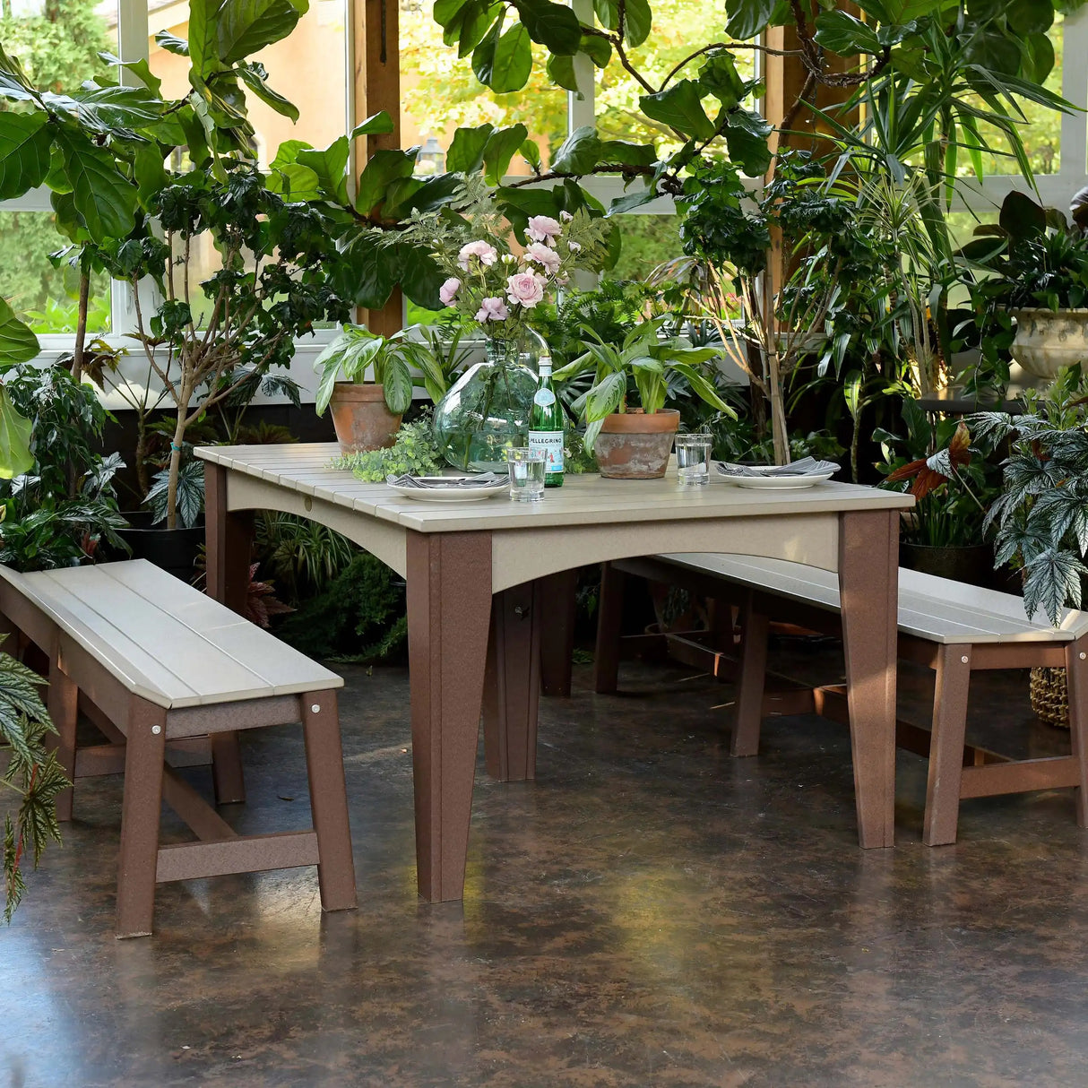 This is a lifestyle image showing the Weatherwood and Chestnut Brown colored dining table with matching benches. This set appears to be in an indoor green room. There are lots of plants around the table and on the table.