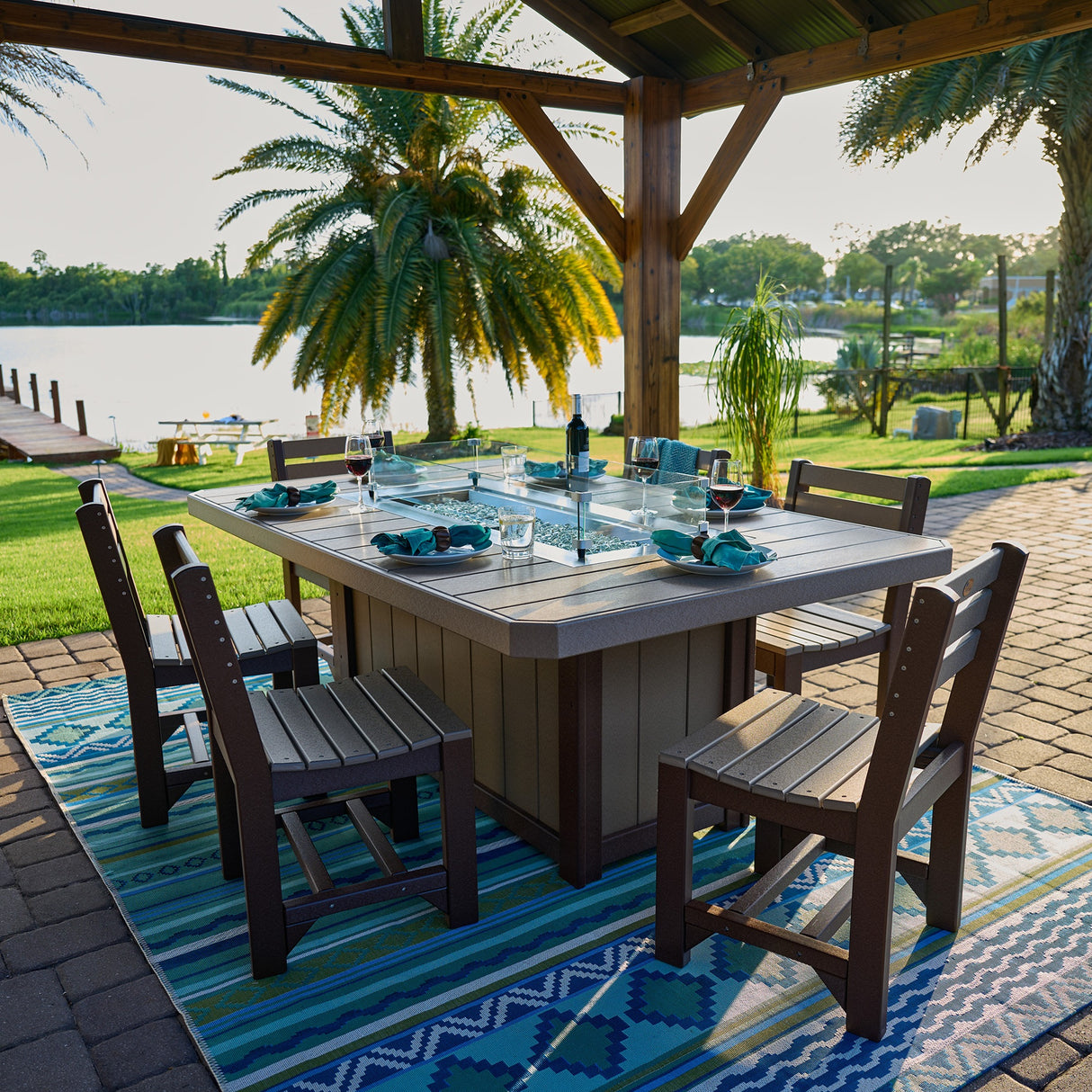This is a lifestyle image showing the Weatherwood and Chestnut Brown colored fire pit table with matching chairs. It's underneath a gazebo in a tropical location.