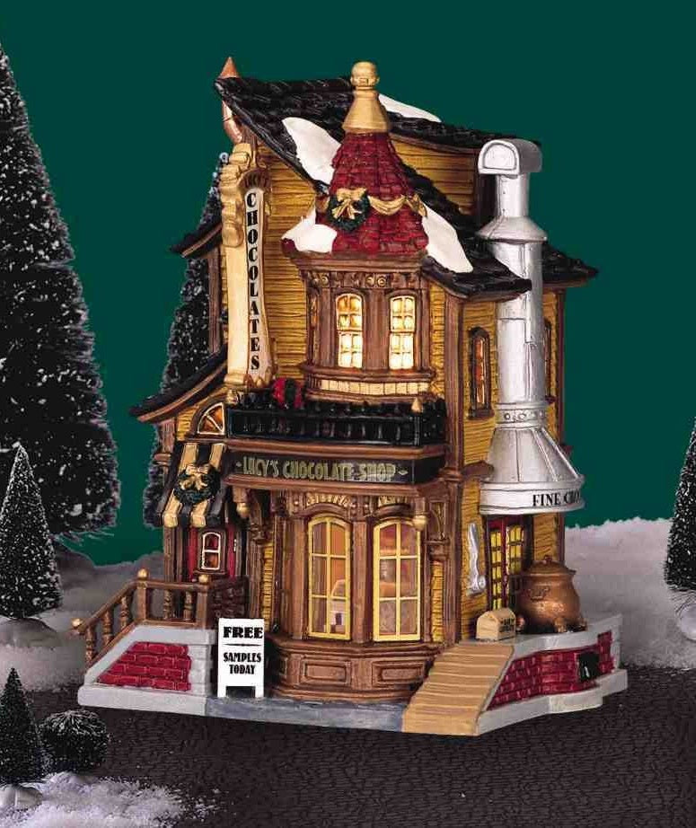 Porcelain lighted building for a Lemax display. Designed to look like an old Victorian-era house with wreaths and snow on it. The house is gold with brown frames. Inside the window you can see an array of chocolates.