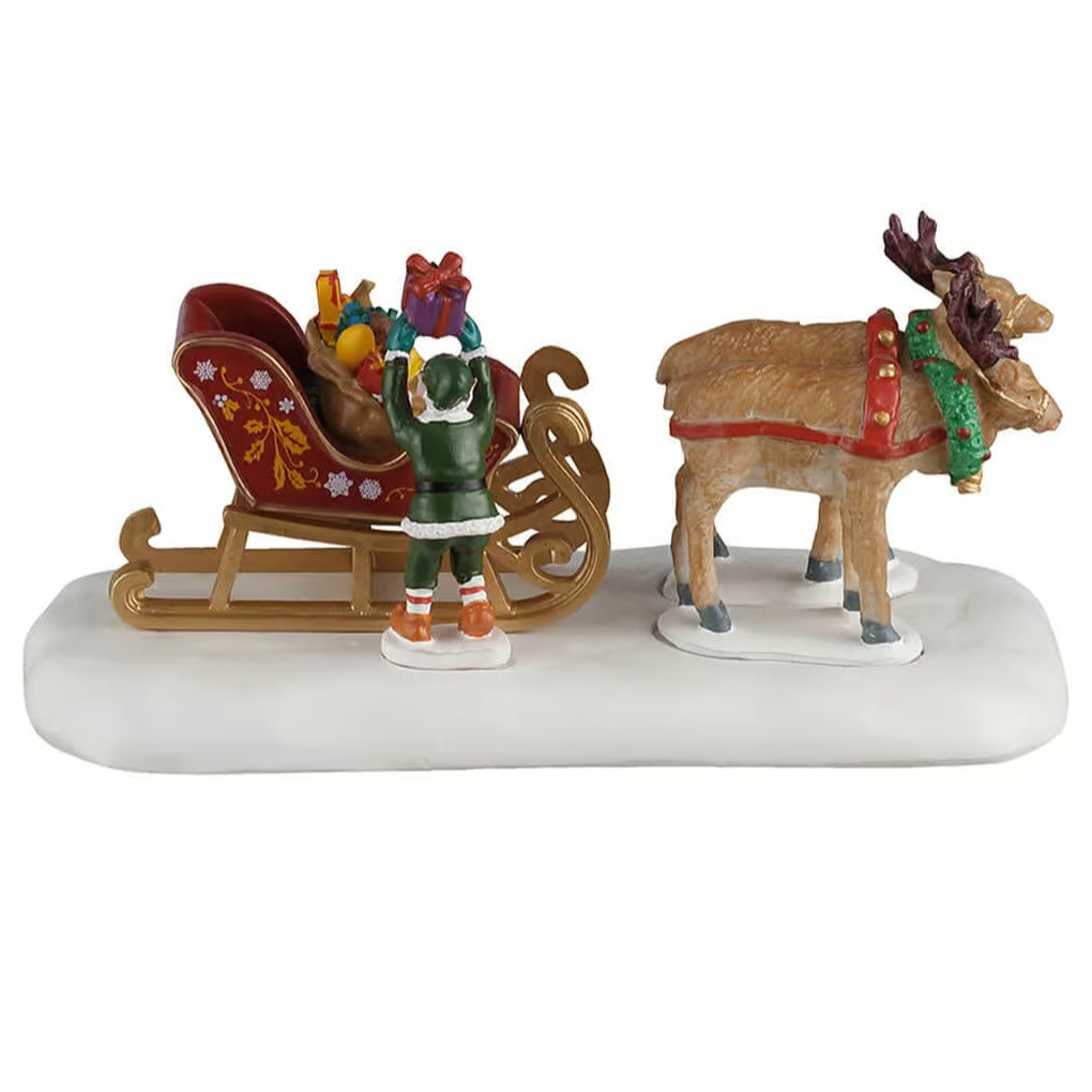 This image shows what this table piece looks like from the side. The elf is wearing a green hat, a white and green jacket, green pants and orange shoes. The sled is red and gold. There's a brown bag of presents in the sleigh. The presents are multi-colored. The reindeer are wearing red reins and green wreaths.