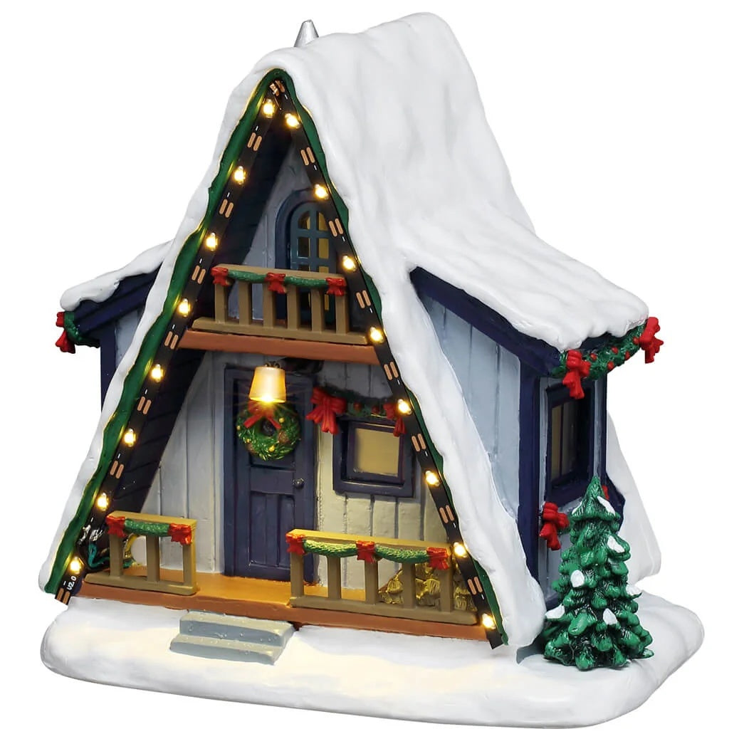 This image shows what this Lemax Vail Village table accent looks like on a white background. This piece was designed to look like an A-frame winter cabin. It's a blue wooden house with lights lining the roofline. The balcony, porch and the sides of the house are all decorated with green garland and red bows. There's a wreath hanging on the door and there's snow all over the house, trees and ground.