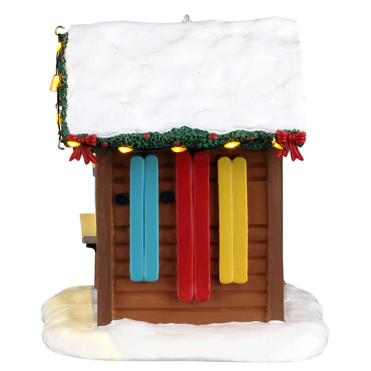This image shows what one of the sides of this building looks like. The roof is covered in snow. There are three pairs of skis hanging on the wall: one set is light blue, one set is red and one set is yellow.