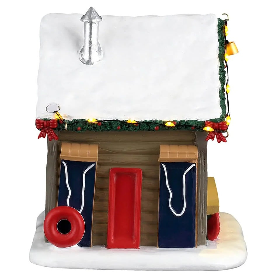 This image shows what one of the sides of the cabin looks like. There's snow on the roof and a silver chimney. The roofline is decorated with lights and a decorated garland. Leaning up against the side, there are three toboggans and one inner tube.