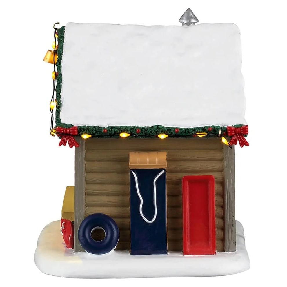This image shows what the other side of the cabin looks like. There's snow on the roof. The roofline is decorated with lights and a decorated garland. Leaning up against the side, there's an inner tube, a toboggan and a sled.