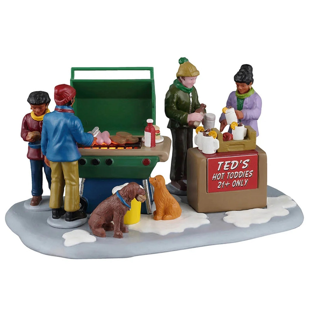 This image shows what this Lemax piece looks like on a white background. It was designed to look like four people grilling together in the snow. There are also two dogs begging for food. There's a sign that reads "Ted's Hot Toddies 21+ Only."
