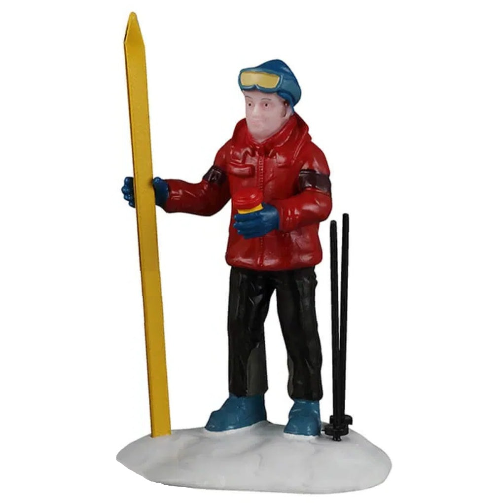 This figurine shows the dad figurine standing in a pile of snow, holding his yellow skis. He's wearing a blue hat, blue goggles, red coat, black pants and blue boots. He's holding a yellow and red colored coffee cup. His black ski poles are standing upright in the snow.