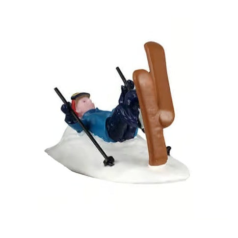 This image shows one of the figurines. It was designed to look like a person who fell backwards while skiing. They're laying on their back in the snow with their skis in the air. They're still holding onto their ski poles. They're wearing a red hat, goggles, a blue jacket and blue pants.